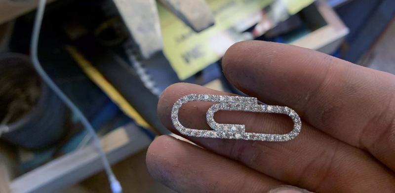 A person holds a diamond-encrusted paperclip-shaped pendant over a jewelry workbench.