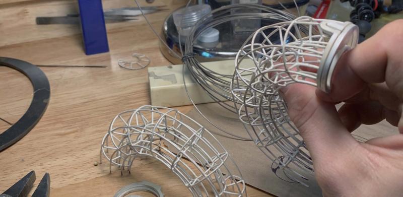 A hand holds a delicate silver wireframe sculpture over a workbench with tools.