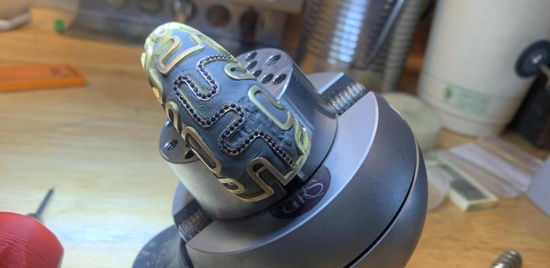 An intricately carved golden bracelet held securely in a silver ball vise on a workbench.