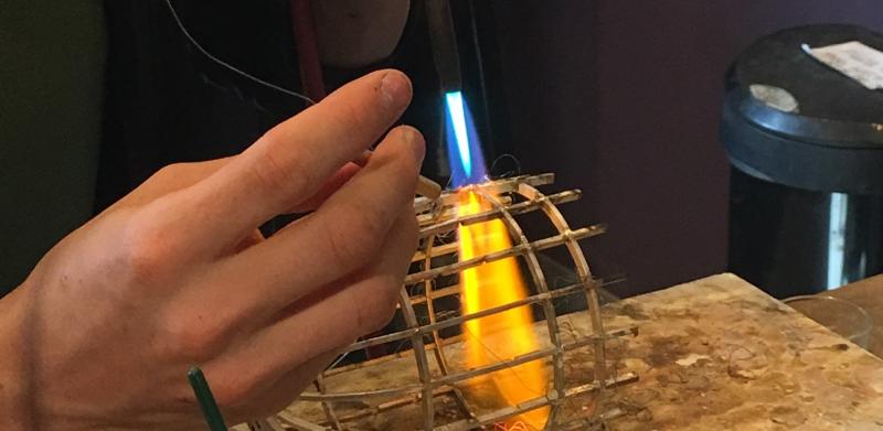A close-up photograph of a person using a blowtorch to solder a metal cage.