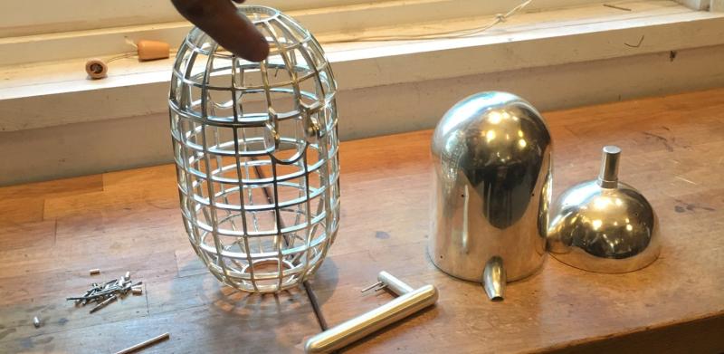 A hand holds a metal mesh cage over a wooden workbench with metal parts.
