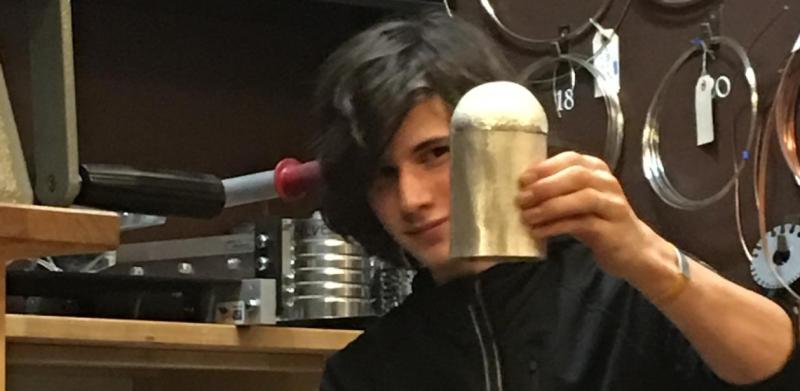 Photograph of a young artisan holding up a metal cylinder in a workshop.