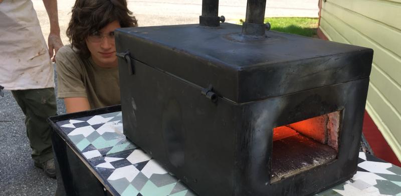 Photo of two people watching a black metal forge on a tiled table outdoors.