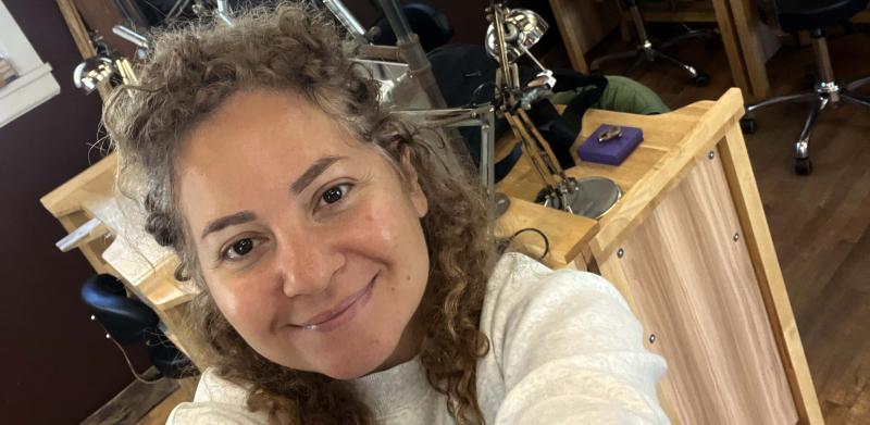 A smiling woman with curly hair takes a selfie in a jewelry workshop.