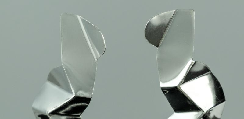 Photograph of two abstract silver metallic geometric earrings on a solid grey background.