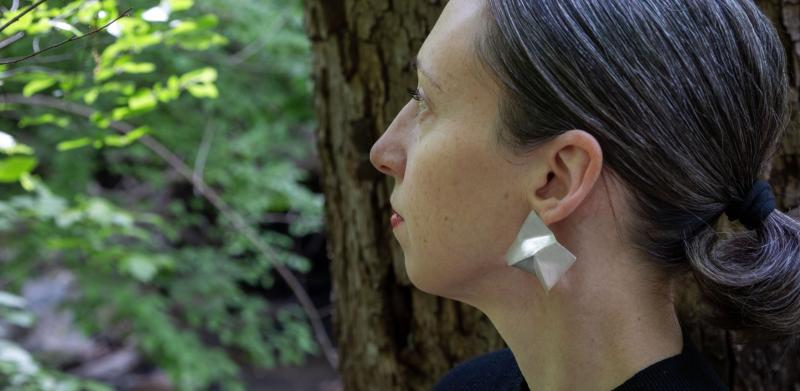 Photograph of a woman wearing silver geometric earrings, standing by a tree in a lush green forest.