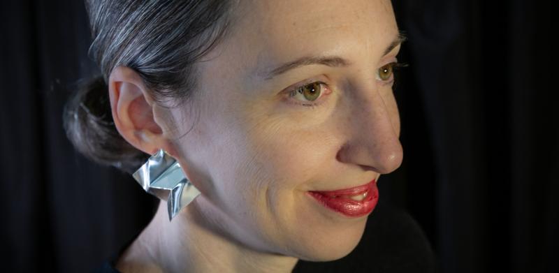 Photograph of a woman with red lipstick and silver origami earrings against a black background.