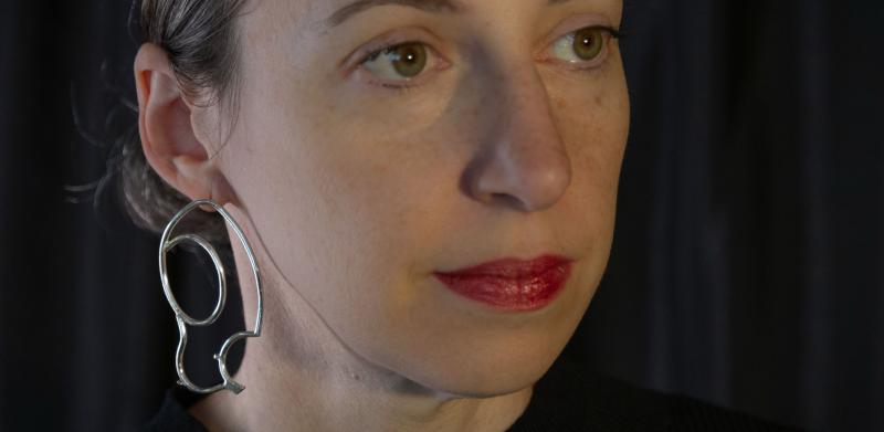 Photograph of a woman wearing large, abstract silver earrings against a dark background.