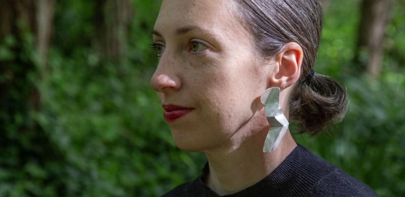 Photograph of a woman with dark hair in a forest wearing a black shirt and silver earrings.
