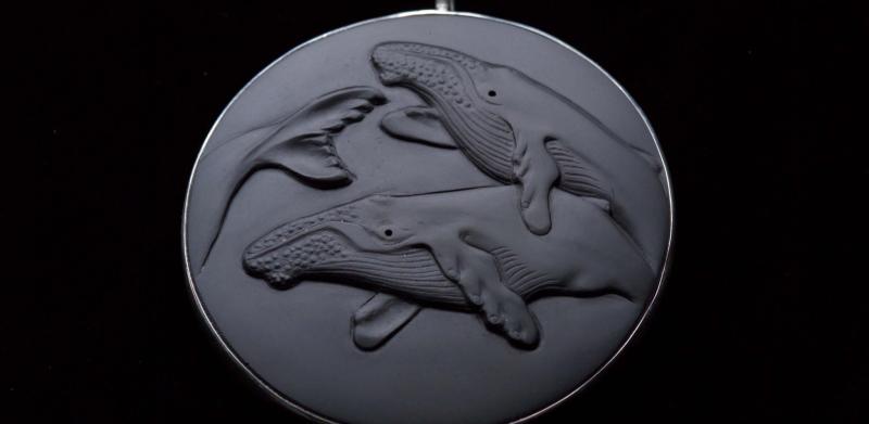 A round dark gray pendant featuring a relief carving of three whales.