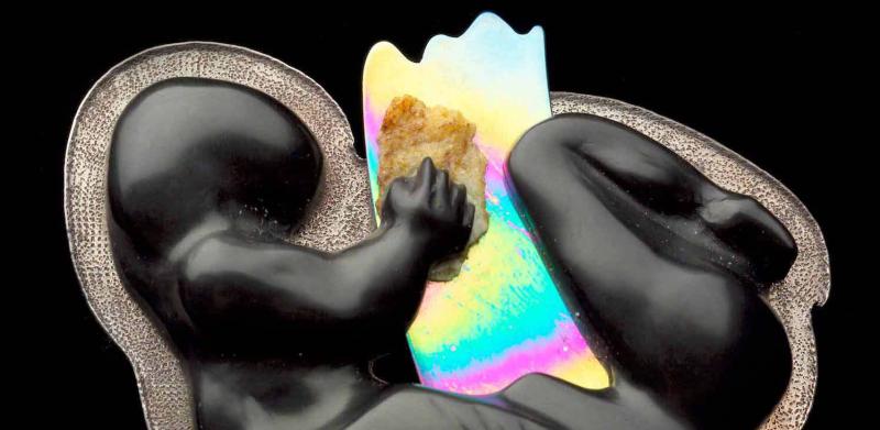 Photograph of a dark, stylized sculpture of a curled fetus holding an iridescent sheet.