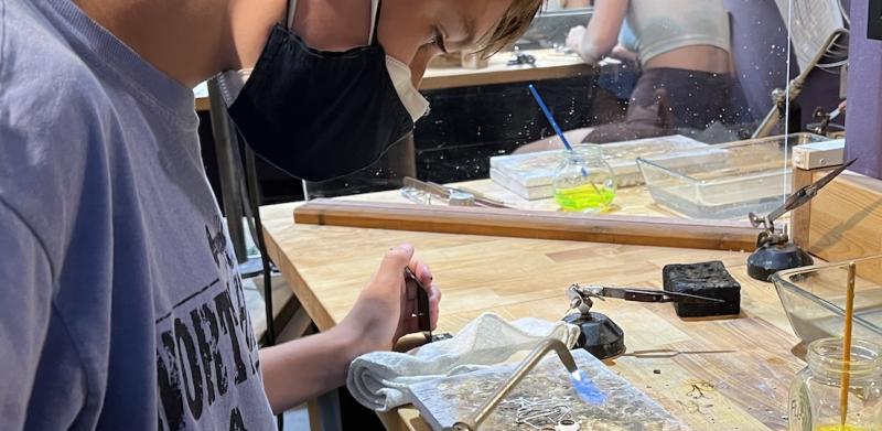 A boy wearing a face mask uses a small blowtorch at a craft table.
