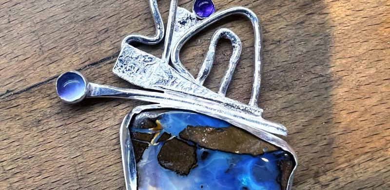 Photograph of an abstract silver pendant with a blue boulder opal and small purple stones.