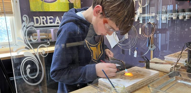 A boy wearing safety goggles uses a torch to heat metal at a workshop table.