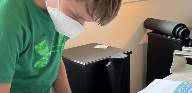 A boy in a green shirt and white mask works on a long banner.