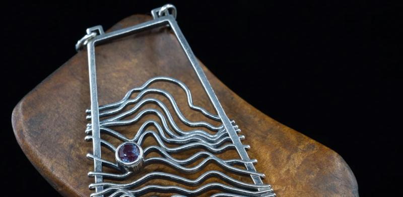 Photograph of a silver wave pendant resting on wood against a black background.