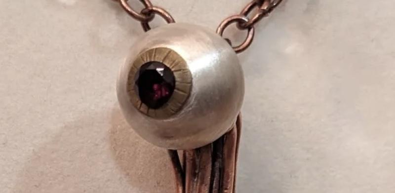 Close-up photograph of a pendant shaped like an eyeball with copper wire tentacles on a chain.