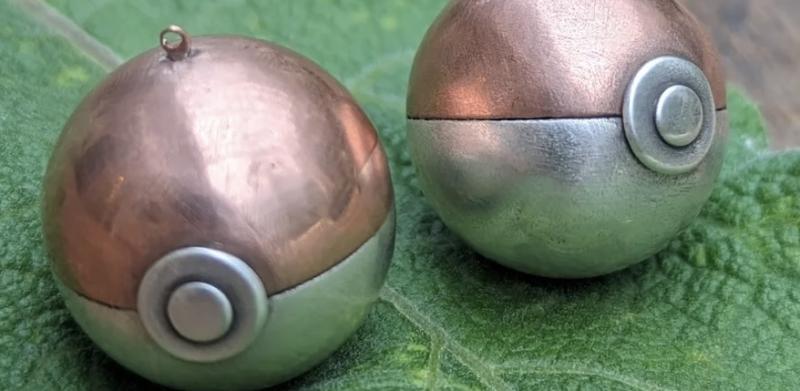 Photograph of two metal Poké Ball pendants resting on a green leaf.