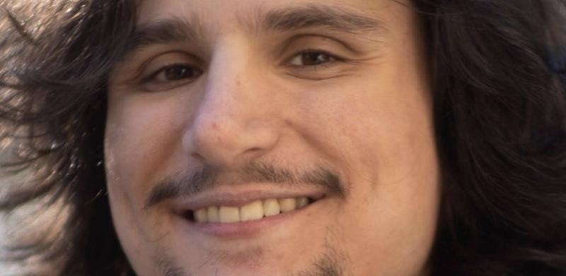Black and white close-up photograph of a smiling young man with long dark hair and a goatee.