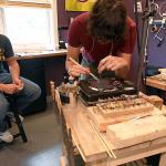 Two people working in a jewelry studio, one focusing on fine detail work at a wooden workbench.