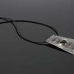 A necklace with a thin, textured, metallic, trapezoid-shaped pendant on a black cord.