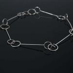 Minimalist silver necklace made of linked thin metal rods and rings on a dark background.