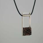 Photograph of a silver, rectangular pendant with a textured dark brown base on a black cord.