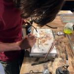 A person wearing a maroon shirt soldering a piece of metal on a wooden workbench.