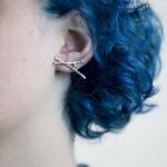 Close-up photograph of a person with blue hair wearing a silver, X-shaped stud earring.