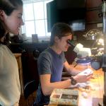 Photograph of a woman standing and watching a man look through a microscope.