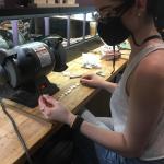 A masked woman grinds pieces of metal at a workbench.