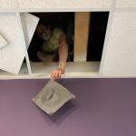 A person crouches in a ceiling opening, holding an air filter.
