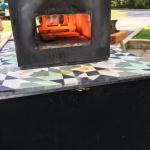 A person's face peers through the glowing, fiery opening of a black metal forge.