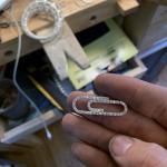A person holds a diamond-encrusted paperclip-shaped pendant over a jewelry workbench.