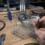 A hand holds a delicate silver wireframe sculpture over a workbench with tools.