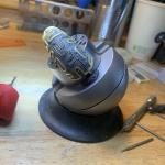 An intricately carved golden bracelet held securely in a silver ball vise on a workbench.