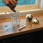 A hand holds a metal mesh cage over a wooden workbench with metal parts.