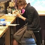 A person with curly brown hair works intently at a workbench in a bright studio.