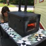 Photo of two people watching a black metal forge on a tiled table outdoors.