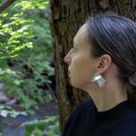 Photograph of a woman wearing silver geometric earrings, standing by a tree in a lush green forest.