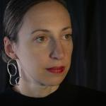 Photograph of a woman wearing large, abstract silver earrings against a dark background.