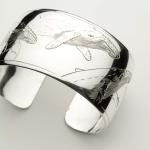 Photograph of a wide silver cuff bracelet engraved with a humpback whale design.