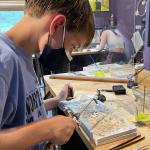 A boy wearing a face mask uses a small blowtorch at a craft table.