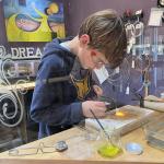 A boy wearing safety goggles uses a torch to heat metal at a workshop table.