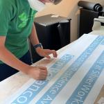 A boy in a green shirt and white mask works on a long banner.