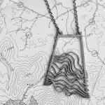 Black and white photo of a trapezoidal pendant necklace on a topographic map.