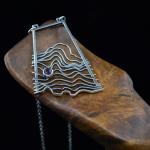 Photograph of a silver wave pendant resting on wood against a black background.