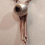 Close-up photograph of a pendant shaped like an eyeball with copper wire tentacles on a chain.