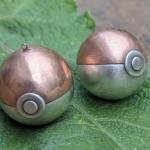 Photograph of two metal Poké Ball pendants resting on a green leaf.